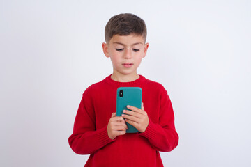 Smiling Little cute boy kid wearing red knitted sweater against white wall using cell phone, messaging, being happy to text with friends, looking at smartphone. Modern technologies and communication.