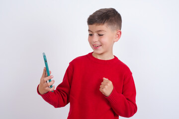 Happy cheerful Little cute boy kid wearing red knitted sweater against white wall receiving good news via e-mail and celebrating success while standing and looking at mobile phone.