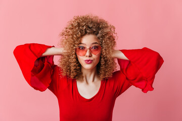 Wall Mural - Close-up portrait of curly blonde in pink sun glasses and red top surprised looking into camera on pink background