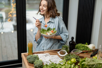 Wall Mural - Portrait of young woman smiling and eating healthy green salad with tomato at home. Health care and wellness concept. High quality photo