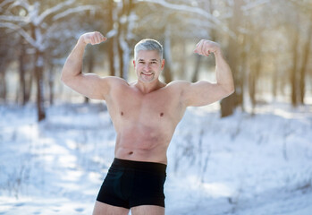 Handsome senior man with bare torso showing his strong biceps in winter frost at snowy forest. Acclimation concept