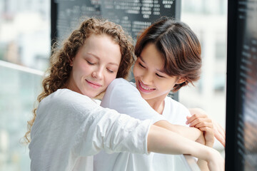 Wall Mural - Happy pretty young curly woman hugging her girlfriend from behind when they are standing on balcony