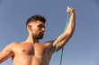 © Andres Victorero/ADDICTIVE STOCK - Muscular male bodybuilder with naked torso standing on seashore and doing exercises with resistance band during workout in summer