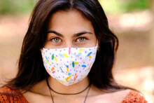 Confident Young Long Haired Brunette In Colorful Cloth Protective Mask For Coronavirus Prevention Looking At Camera While Standing Against Blurred Background Of Autumnal Park