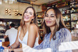 © David Espejo/ADDICTIVE STOCK - Smiling young girlfriends looking at camera while sitting at table with drinks during meeting in modern cafeteria in summer day