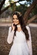© German Linares/ADDICTIVE STOCK - Charming tender young brunette in stylish white lace wedding dress and bridal wreath touching cheek and smiling while standing near tree in park