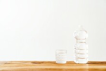 Empty Glass And Plastic Bottle Filled With Pure Fresh Water Placed On Wooden Table Against White Wall