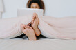 © Jake Jakab/ADDICTIVE STOCK - Young ethnic barefoot female covered with blanket sitting in bed and browsing laptop at home