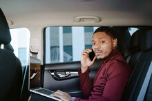 Side View Of Busy African American Businessman Sitting In Luxury Car With Laptop And Speaking With Client On Smartphone While Looking At Camera