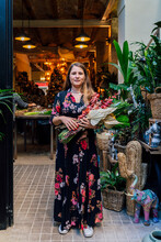 Woman Holding Flower Bouquet Standing Outside Home Decor Shop In Daylight Looking At Camera