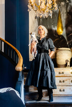 Full Body Of Confident Stylish Middle Aged Female In Elegant Black Dress Holding Little Dog And Looking At Camera While Standing Near Stairs In Room With Vintage Furniture And Chandelier At Home