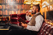 © Javier De La Torre/ADDICTIVE STOCK - Side view of relaxed bearded male using tablet and enjoying liquor while resting on sofa in elegant pub