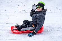 Happy Boy Wearing Sunglasses Ride From A Snow Slide On A Red Pan Sledge. Winter Activities. Downhill. Sledging. Winter Entertainment And Sports. Activities For Children And Teenagers. Speed Sled - Ice