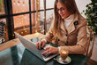 © Jesus Diaz/ADDICTIVE STOCK - Smiling young female blogger in casual clothes and glasses sitting at table with cup of coffee and typing on laptop keyboard while preparing content for social networks in cozy cafeteria