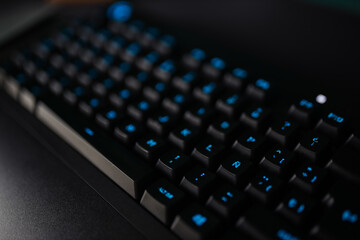 From above of black keyboard with blue illuminated letters placed on desk near computer monitor