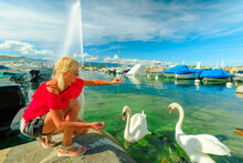 Blonde Tourist Girl Feeding White Swans In Turquoise Waters Of Geneva Lake In Geneva Harbor With 140m High Fountain Called Jet D'Eau. Marina With Boats, Sailboats And Yachts. French Swiss, Switzerland