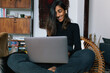 © Joy Zamora/ADDICTIVE STOCK - Cheerful Indian female sitting in wicker chair and having video call on laptop while smiling