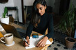 © Joy Zamora/ADDICTIVE STOCK - High angle of Indian female entrepreneur sitting at table and writing plans in notebook while working at home in cozy workplace