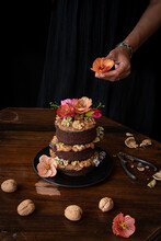 Crop Anonymous Chef Decorating Festive Rustic Chocolate Naked Cake With Walnuts And Natural Edible Flowers