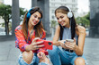 © Julio Rodriguez/ADDICTIVE STOCK - Cheerful hipster female students in trendy outfits surfing social networks on mobile phones and listening to music with headphones while sitting together on urban street in summer day