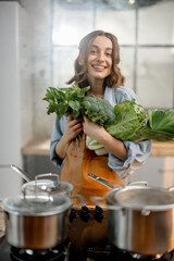 Pretty housewife in apron with fresh vegetables near cooker with boiling pans on the kitchen. Healthy green cooking concept. High quality photo