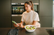 © Luis Tenza/ADDICTIVE STOCK - Peaceful female sitting at table in kitchen and eating tasty vegetable salad while reading news on social media on smartphone