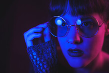 Tranquil Female In Stylish Round Glasses Standing On Dark Background In Pink And Blue Neon Illumination