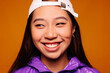 © Victor Torres/ADDICTIVE STOCK - Portrait of happy Asian young woman. She wears a purple jacket and a grey cap and is looking away smiling against a yellow background