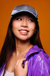 © Victor Torres/ADDICTIVE STOCK - Portrait of happy Asian young woman. She wears a purple jacket and a grey cap and is looking away smiling against a yellow background