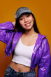 © Victor Torres/ADDICTIVE STOCK - Portrait of happy Asian young woman. She wears a purple jacket and a grey cap and is looking at camera smiling against a yellow background