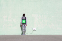 Back View Of Unrecognizable Female With Long Dark Hair In Stylish Outfit Standing On Street Near Wall With Battery Shaped Backpack Connected To Charger
