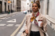 © The Trio Studio/ADDICTIVE STOCK - Confident young African American female entrepreneur in fashionable outfit listening to music with wireless earphones and drinking takeaway coffee while walking on city street during break