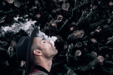 Side View Of Male Hipster In Hat And Red Shirt Exhaling Cloud Of Smoke Standing Along With Eyes Closed Against Pile Of Logs In Nature