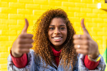 Optimistic Young Black Female Millennial With Curly Hair In Stylish Clothes Showing Thumbs Up Gesture And Smiling While Looking At Camera Against Yellow Wall