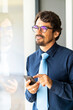 © Joao - Businessman wearing glasses smiling, holding his mobile phone and looking through the window. Happy successful male portrait