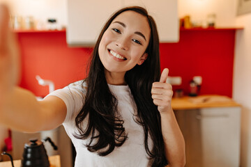 long-haired woman dressed in white t-shirt makes selfie in kitchen and shows thumb up