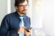 © Joao - Business man wearing glasses looking through window holding his smartphone. Successful male portrait with serious face at the office by the window