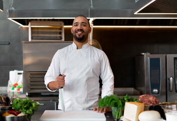 Wall Mural - Handsome young African chef standing in professional kitchen in restaurant preparing a meal of meat and cheese vegetables.