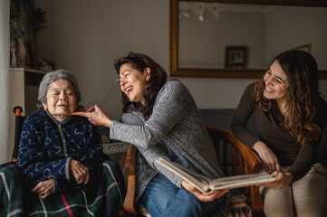 women generation with old sick grandmother sitting in wheelchair and smiling daughter and granddaughter looking a photo album