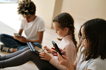 Three multiethnic little kids sit indoor using digital gadgets