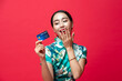 © Atstock Productions - Surprised Asian woman wearing oriental qipao dress looking at credit card in hand on red studio background for Chinese new year cashless shopping concepts