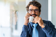 © Joao - Business man wearing glasses, smiling while talking on the phone and drinking coffee. Happy successful male portrait at the office by the window