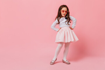 Charming girl in fluffy skirt posing on pink background. Curly baby wearing heart-shaped glasses