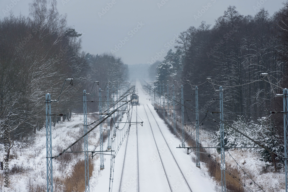 Dwutorowy szlak kolejowy zimą z przejeżdżającą lokomotywą elektryczną. - obrazy, fototapety, plakaty 