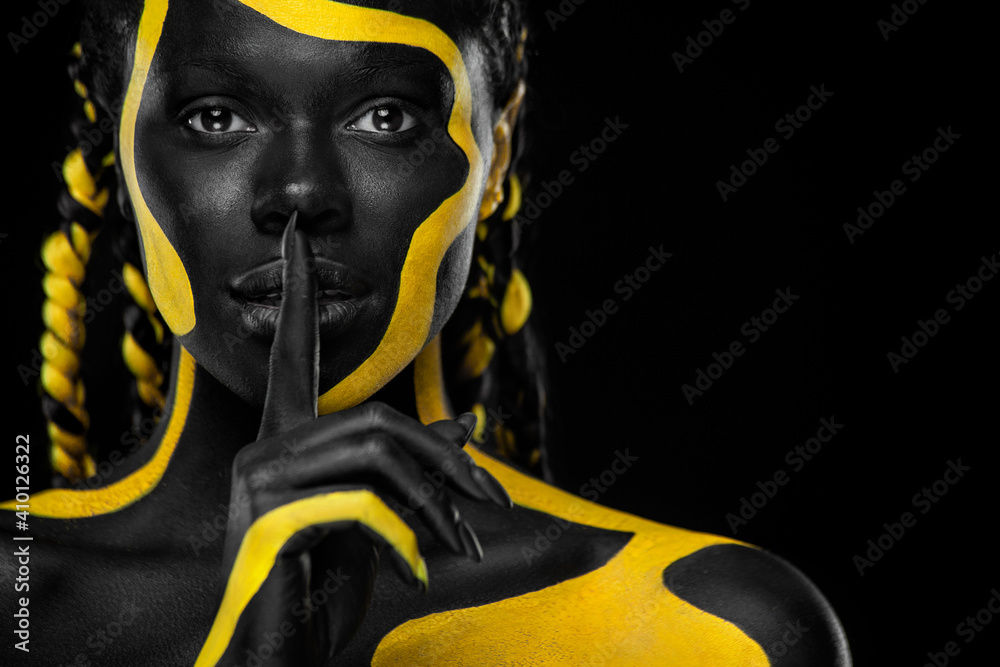 Face art. Girl showing gesture quietly, asks remain silent with finger. Woman with black and yellow body paint. Young african girl with colorful bodypaint. An amazing afro american model with makeup. - obrazy, fototapety, plakaty 