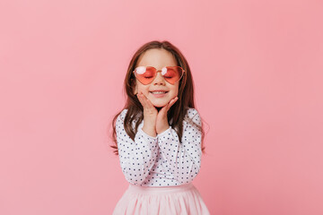 cute female child in light outfit and pink sunglasses smiles with closed eyes on pink background
