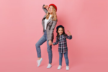 Wall Mural - Adult woman and female child pose against pink background and show peace sign. Portrait of mother and daughter in plaid shirts and berets