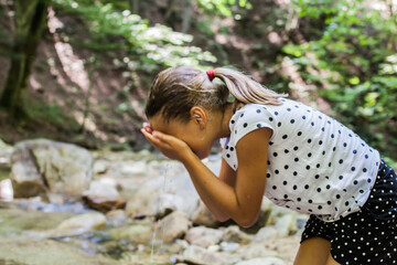 Wall Mural - Little girl refreshing by clear water from forest mountain creek on summer day in beautiful nature.