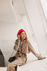 Wall Mural - Slim lady in elegant beige trench coat and red beret touches her hair and sits by window on white background