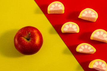 red apple stands on a yellow background next to pink and white marshmallows on a red background. creative idea of choosing between useful and harmful. fruits and high-calorie foods.